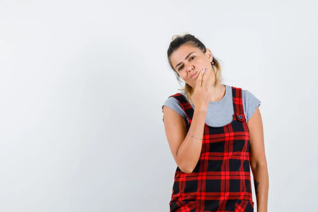 Young girl with a thoughtful expression, touching her chin, wearing a red and black plaid dress over a gray shirt, against a plain white background, symbolizing contemplation related to oral health concerns like gum recession.