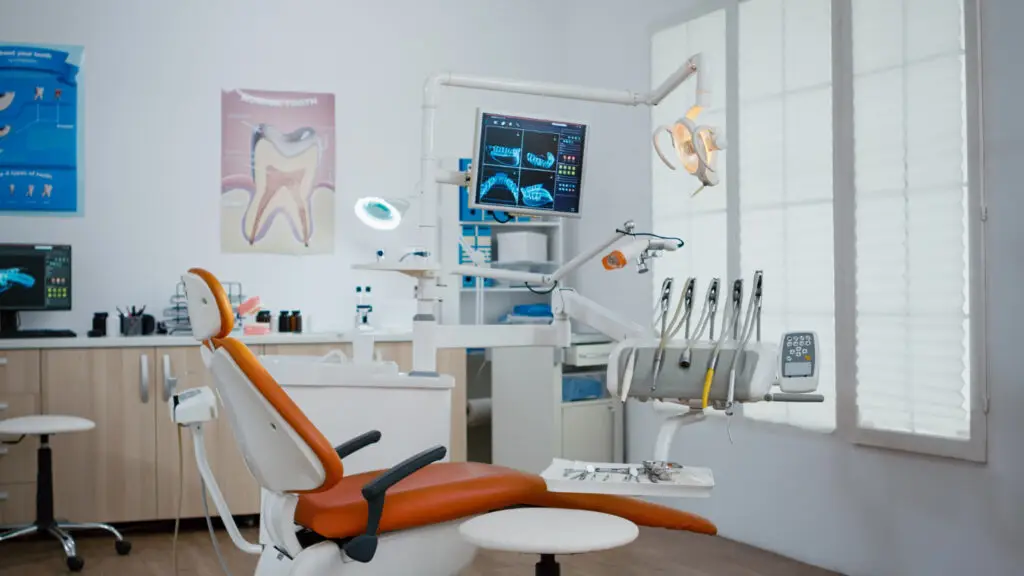 Modern dental treatment room with orange patient chair, digital X-ray monitor, dental tools, and exam lighting for care.